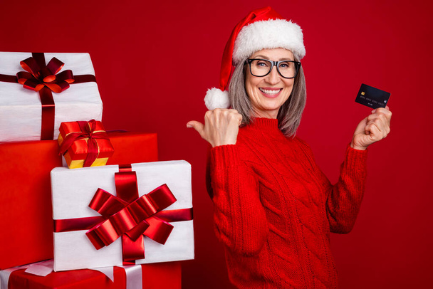 Photo of grandma hold credit card direct finger pile gift wear specs hat pullover isolated red color background. - Foto, afbeelding