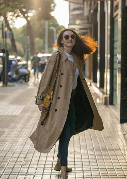 Felnőtt 35 éves üzletasszony hosszú hajjal, napszemüveggel és ballonkabáttal séta egy modern épületekkel rendelkező városban. Városi jelenet Barcelona város. - Fotó, kép