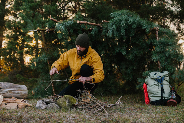 Sopravvivenza in natura. Un uomo barbuto accende un fuoco vicino a un rifugio improvvisato fatto di rami di pino. - Foto, immagini