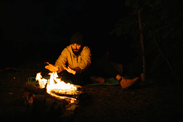 El viajero descansa junto a la fogata nocturna. Amueblado refugio de ramas para dormir con una lámpara de queroseno y un fuego rodeado de piedras. - Foto, Imagen