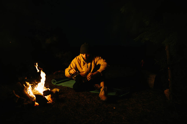 Il viaggiatore riposa vicino al falò notturno. Rifugio arredato da rami per dormire con una lampada al cherosene e un fuoco circondato da pietre. - Foto, immagini