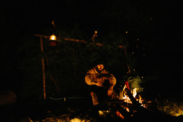 El viajero descansa junto a la fogata nocturna. Amueblado refugio de ramas para dormir con una lámpara de queroseno y un fuego rodeado de piedras. - Foto, Imagen