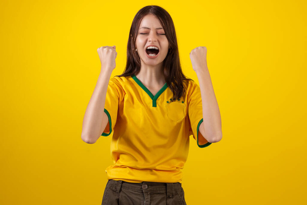 A nő a brazil focicsapat hivatalos egyenruháját viseli a 2022-es Katar Kupán, és Brazília zászlaját viseli a stúdiófotón. Brazil ventilátor. - Fotó, kép
