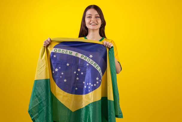 oung γυναίκα φορώντας το επίσημο πουκάμισο στολή της ομάδας ποδοσφαίρου της Βραζιλίας στο 2022 Κύπελλο Κατάρ και με τη σημαία της Βραζιλίας στη φωτογραφία στούντιο. Βραζιλιάνος οπαδός. - Φωτογραφία, εικόνα