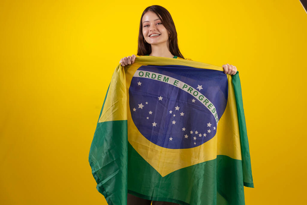 oung γυναίκα φορώντας το επίσημο πουκάμισο στολή της ομάδας ποδοσφαίρου της Βραζιλίας στο 2022 Κύπελλο Κατάρ και με τη σημαία της Βραζιλίας στη φωτογραφία στούντιο. Βραζιλιάνος οπαδός. - Φωτογραφία, εικόνα