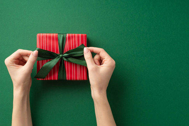 Η ιδέα της παραμονής Χριστουγέννων. Φωτογραφία πρώτου προσώπου με νέα γυναικεία χέρια που δένουν κορδέλα σε κόκκινο κουτί δώρου σε απομονωμένο πράσινο φόντο με κενό χώρο - Φωτογραφία, εικόνα