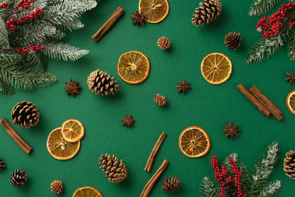 Christmas decorations concept. Top view photo of dried orange slices anise cinnamon sticks pine cones mistletoe berries and fir branches in snow on isolated green background - Photo, Image