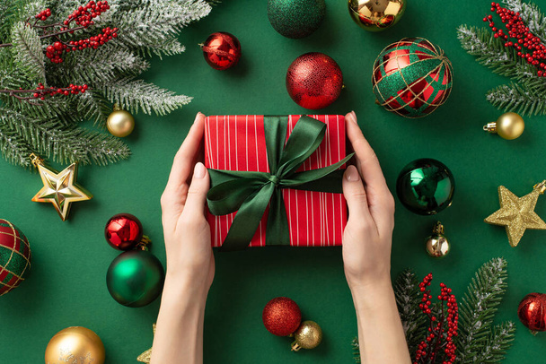 Nieuwjaarsconcept. Eerste persoon bovenaanzicht foto van meisje handen houden rode giftbox met boog goud groene kerstballen ster ornamenten maretak bessen sparren takken in de vorst op geïsoleerde groene achtergrond - Foto, afbeelding