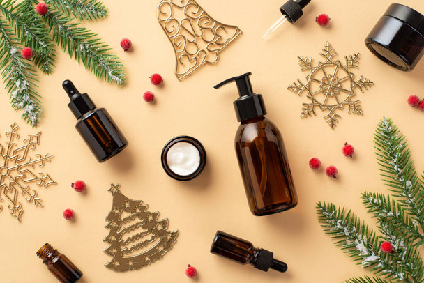 Winter skin care concept. Top view photo of amber glass bottles without label cream jar christmas tree ornaments mistletoe berries fir branches in frost on isolated beige background with empty space - Photo, image