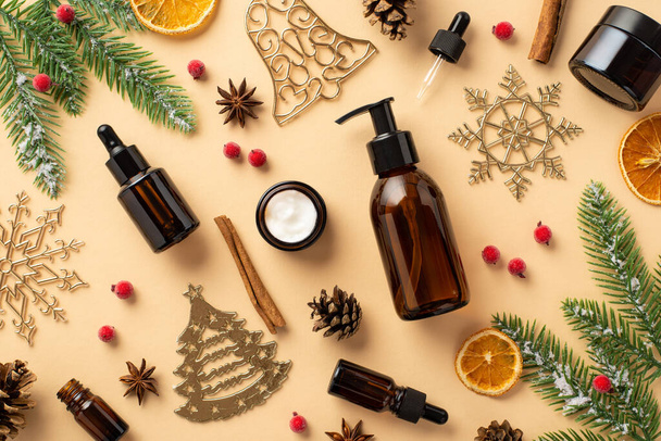Winter skincare cosmetics concept. Top view photo of pump bottle dropper bottle cream jar christmas ornaments mistletoe fir branches pine cone cinnamon dried orange slices on isolated beige background - Photo, Image