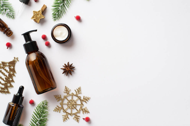 Winter season skin care concept. Top view photo of amber glass bottles cream jar christmas decor golden ornaments fir branches and mistletoe berries on isolated white background with empty space - Photo, Image