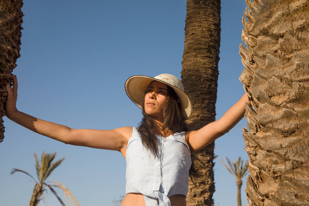 Sexy turista entre las palmeras buscando y disfrutando del desierto del palmeral de Marrakech en Marruecos con un sombrero, este es un lugar muy visitado por los turistas de vacaciones y diversión. - Foto, imagen