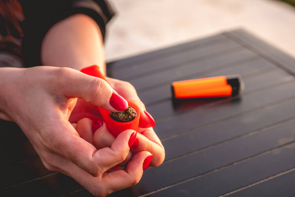 Mani con chiodi rossi mettere un germoglio d'erba in un tubo di plastica di marijuana arancione e accendino arancione con bella luce del tramonto - Foto, immagini