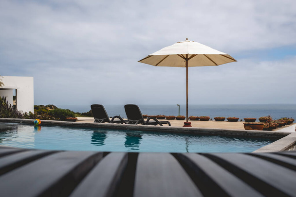 Καρέκλα παραλίας μπροστά από μια μεγάλη πισίνα στη βεράντα με περισσότερες καρέκλες, ομπρέλα και τον ωκεανό σε ένα καλοκαιρινό βράδυ - Φωτογραφία, εικόνα