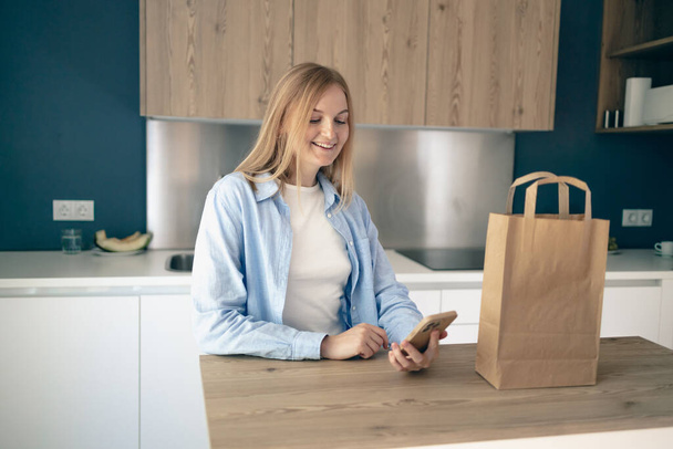 Young lady unpacking paper bag after shopping using smartphone in kitchen interior at home, copy space. - Photo, Image