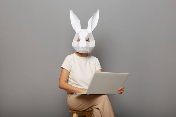 Portrait of anonymous female wearing rabbit mask and white t-shirt, sitting on chair with notebook in hands, freelancer working online, isolated over gray background. - Photo, Image