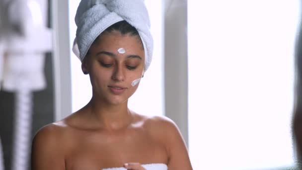 Donna dopo una doccia con un asciugamano sulla testa applica una crema idratante per il viso, le procedure quotidiane del mattino la cura della pelle. - Filmati, video