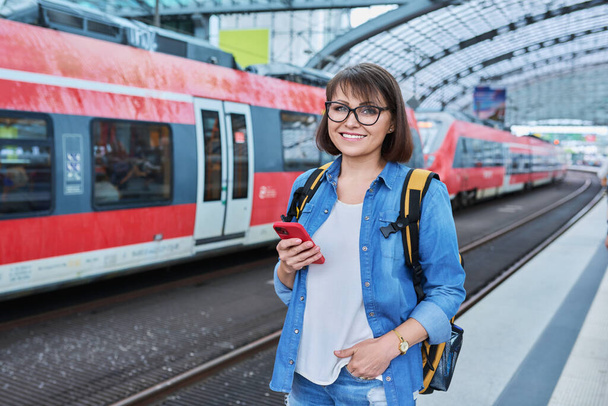 Woman waiting for railway public electric transport on platform of city station. Female with smartphone using banking app to buy online ticket payment, online timetable and route service, technology - Photo, image