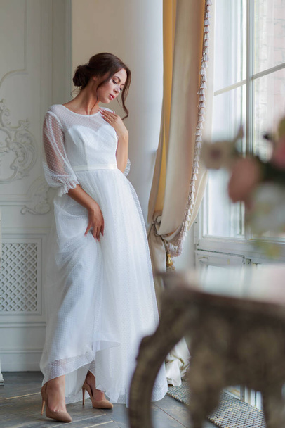Full-length portret van de bruid in een witte trouwjurk bij het raam  - Foto, afbeelding