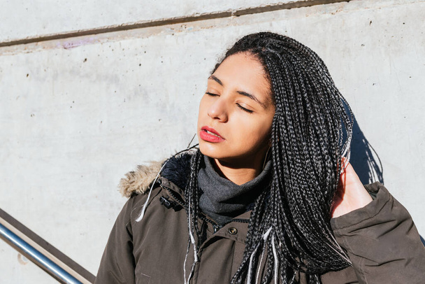 Tranquil female with braids touching black hair and enjoying sunny weather with closed eyes in street - Zdjęcie, obraz