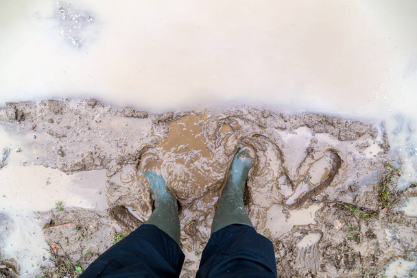 stopy w zielonych gumowych butach stoi w mokrej brązowej kałuży błota bezpośrednio nad widokiem - Zdjęcie, obraz