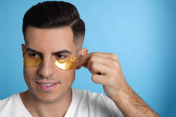 Man applying golden under eye patch on light blue background - Foto, Bild