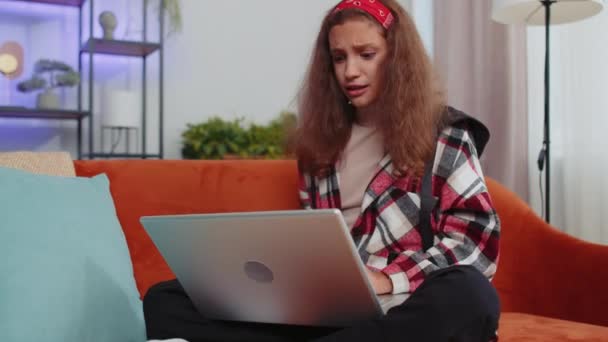 Portret van Kaukasische tiener meisje gebruiken laptop typen browsen werken, verliest steeds verrast plotselinge loterij resultaten, slecht nieuws, fortuin verlies, falen. Jong kind, kind thuis in kamer op oranje bank - Video