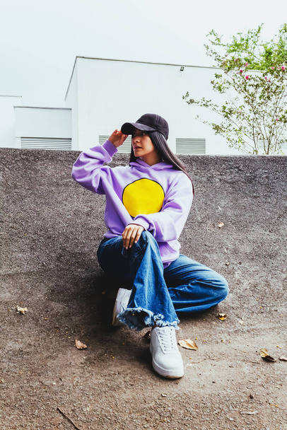 belle adolescente en sweat-shirt et casquette assis dans la ville dans un style urbain posant à la caméra, heureuse fille latine à la mode en plein air - Photo, image