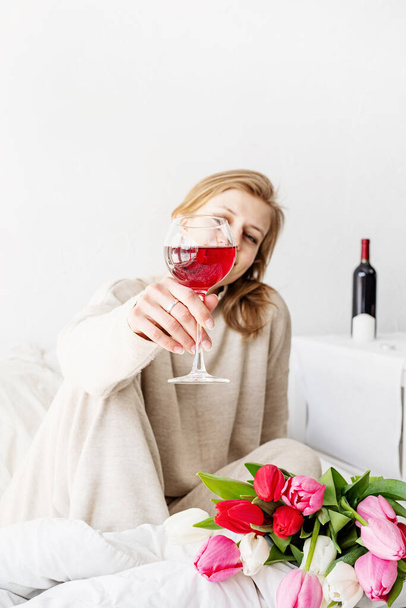Mutlu kadın pijamalarıyla yatakta oturuyor. Elinde bir bardak şarap ve lale çiçeği buketi var. - Fotoğraf, Görsel