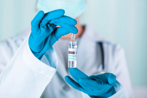 "Врач или медсестра в форме и перчатках в защитной маске для лица в лаборатории держат пузырек с лекарственной вакциной с этикеткой короновирусной вакцины COVID-19" - Фото, изображение