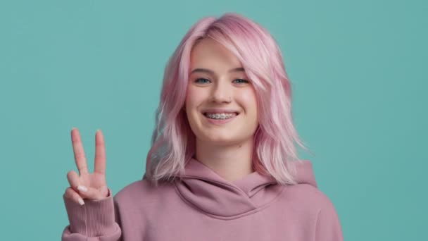 Χαρούμενο χαμογελαστό χαρούμενο κορίτσι με ροζ μαλλιά Gesturing V-Sign Smiling To Camera Posing Over Teal Blue Studio Background. Casual Fashionable Γυναίκα δείχνει Νίκη χειρονομία. Έννοια θετικής κινητοποίησης - Πλάνα, βίντεο