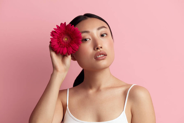 Skin Care Woman Posing with Red Flower. Asian Girl Holding Flower and Looking At Camera. Female With Nude Makeup, Smooth, Hydrated And Glowing Skin. Natural Beauty On Pink Background  - Fotoğraf, Görsel