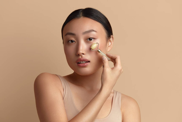 Face massage. Asian Woman Using Jade Facial Roller for Skin Care, Beauty Treatment on Beige Isolate Background. Girl Using Natural Massager Closeup Portrait  - Photo, Image