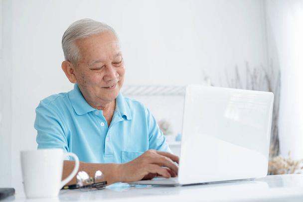 Счастливый пожилой человек, работающий на ноутбуке и держащий чашку кофе - Фото, изображение