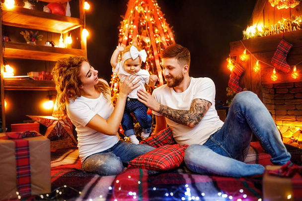Happy mother and father playing with cute baby daughter while sitting together near Christmas tree and fireplace in decorated living room. New Year concept  - Foto, imagen