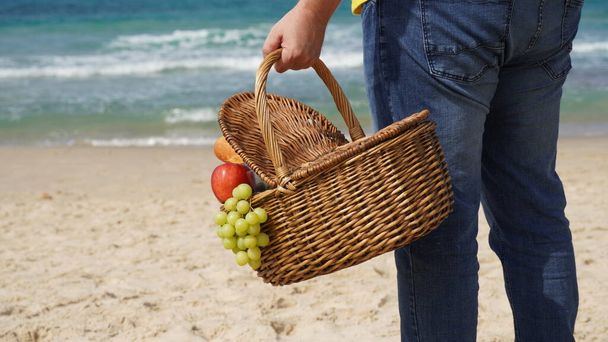 A man holds a wicker basket with food and wine for picnic on the beach. Summertime relaxation and recreation concept. - Foto, imagen