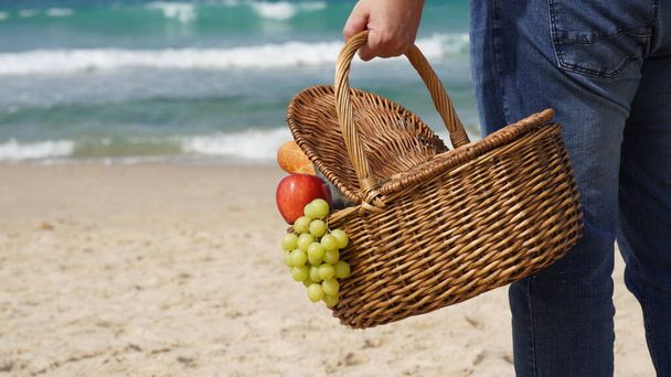 A man holds a wicker basket with food and wine for picnic on the beach. Summertime relaxation and recreation concept. - Foto, Imagem