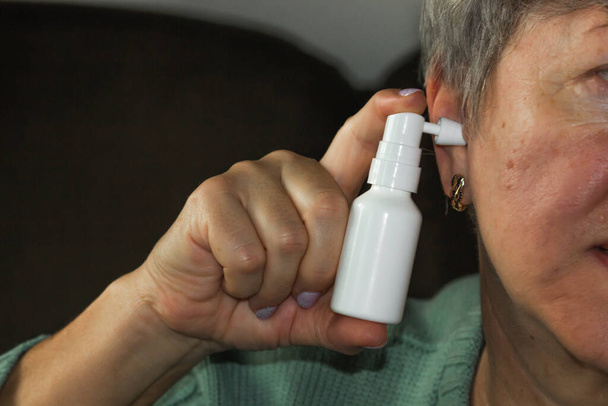 Старшая белая женщина использует спрей для ушей. Женские руки держат в руках белый баллончик с насадкой в ушной раковине. Ежедневная гигиена и чистка ушей от воска. Концепция здравоохранения. - Фото, изображение