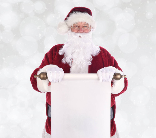 サンタクロースは、雪の効果とボケの背景の上に、彼の体の前に紙のスクロールを保持する。紙はあなたのコピーのための部屋と空白です - 写真・画像