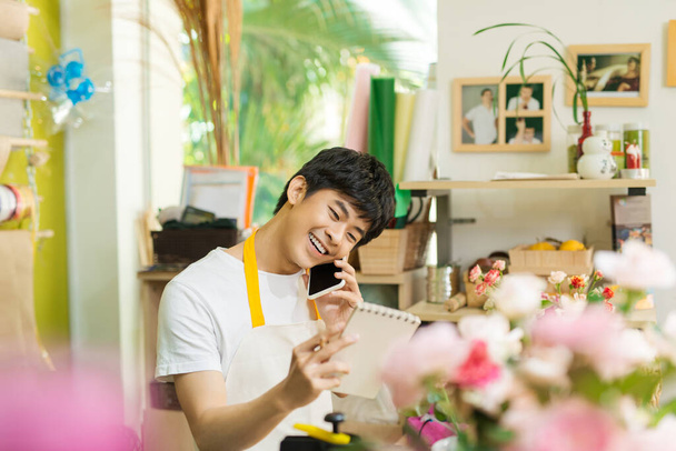 "Sorrindo asiático florista masculino chamando no smartphone e fazendo notas no balcão da loja de flores" - Foto, Imagem
