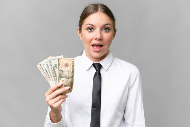Joven mujer caucásica de negocios sosteniendo el dinero sobre fondo aislado con sorpresa y expresión facial impactada - Foto, imagen
