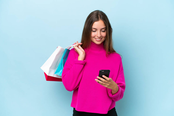 Jonge mooie vrouw geïsoleerd op blauwe achtergrond met winkeltassen en het schrijven van een bericht met haar mobiele telefoon naar een vriend - Foto, afbeelding