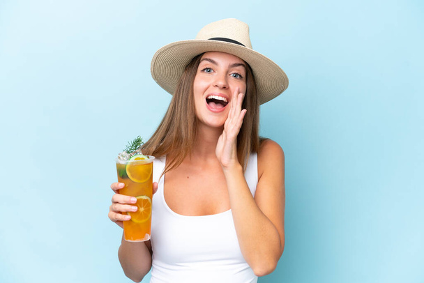 Junge schöne Frau hält einen Cocktail isoliert auf blauem Hintergrund und schreit mit weit geöffnetem Mund - Foto, Bild