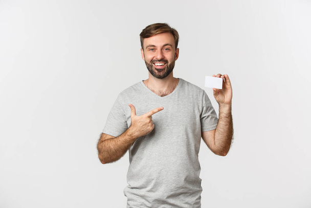 Portrét pohledného vousatého muže v šedém tričku, ukazuje na kreditní kartu a usmívá se, stojí nad bílým pozadím - Fotografie, Obrázek