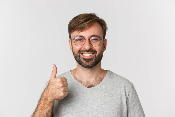 Крупный план жизнерадостного кавказца с бородой, в очках и повседневной футболке, улыбающегося и показывающего большие пальцы на одобрение, рекомендую продукт, стоя на белом фоне - Фото, изображение