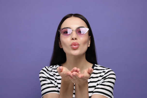 Hermosa mujer joven en gafas de sol con estilo soplado beso sobre fondo púrpura - Foto, imagen