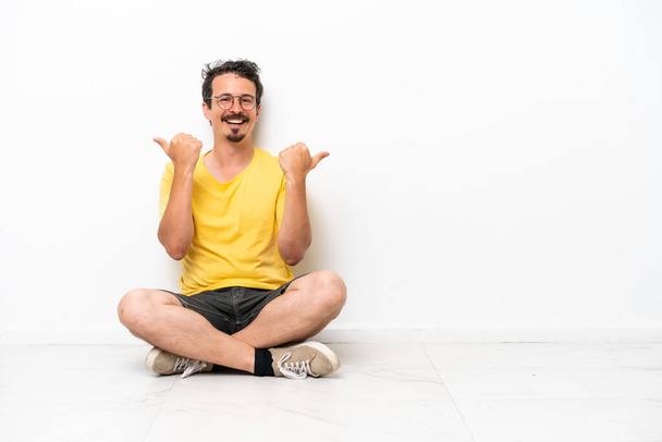 Joven hombre caucásico sentado en el suelo aislado sobre fondo blanco con los pulgares hacia arriba gesto y sonriendo - Foto, imagen
