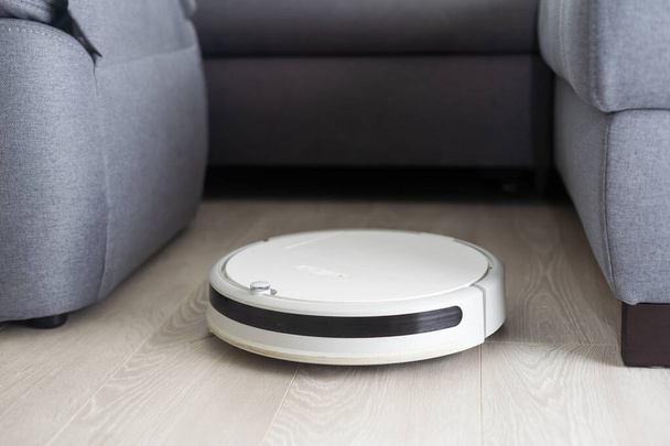 A white robot vacuum cleaner vacuums along the skirting Board near a large window through which the bright sun shines. close-up, beige colors, small depth of field. - Foto, immagini