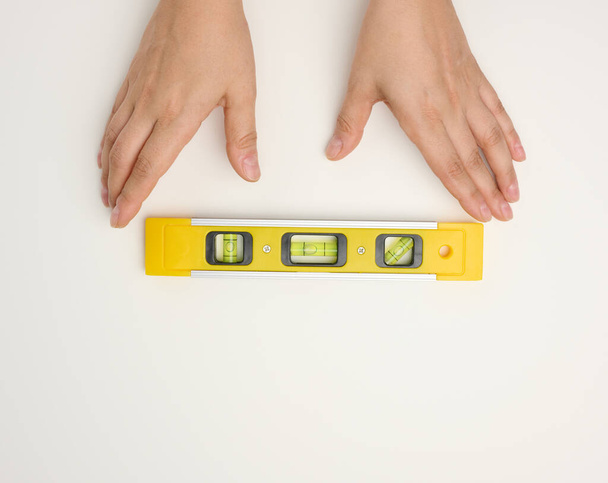manos femeninas sostiene un nivel de construcción de plástico amarillo sobre un fondo blanco - Foto, imagen