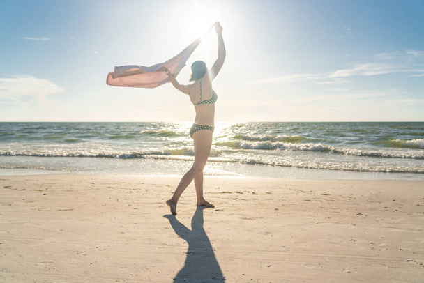 Mulher de pé com toalha na praia com oceano no fundo
 - Foto, Imagem